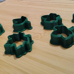 May include: Several dark green, clover-shaped cookie cutters are arranged on a light brown wooden surface. The cutters have a three-leaf design, perfect for making St. Patrick's Day treats or other themed cookies. The cutters are made of plastic.