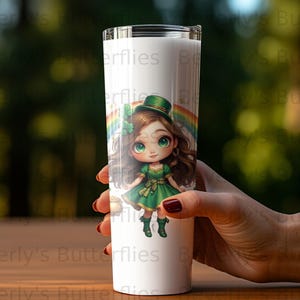May include: A white tumbler featuring a cartoon leprechaun girl with a green dress, hat, and boots, and a rainbow background. The tumbler is held by a hand.