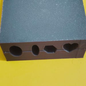 May include: A dark gray and brown rectangular mold with four differently shaped cutouts: a circle, an oval, a hexagon, and a heart. The mold has a textured surface and sits on a yellow background.