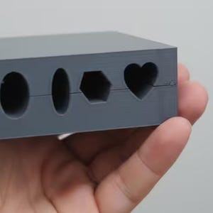 May include: A gray rectangular mold featuring four distinct cutouts along its side. The openings include a large circle, a smaller oval, a hexagon, and a heart shape, all aligned horizontally across the center of the block.