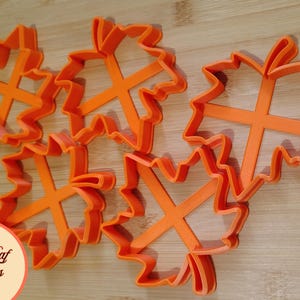 Könnte beinhalten: Orangefarbene Ausstecher in Ahornblattform aus Kunststoff auf einer Holzoberfläche. Jeder Ausstecher hat eine X-förmige Stütze. Der Text "Fall Leaf Cutters" befindet sich in einem Kreis unten links.