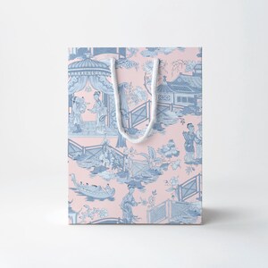 Op de afbeelding: Een roze cadeauzakje met een blauw en wit Aziatisch geïnspireerd toile-patroon. De tas heeft een herhalend ontwerp van figuren, gebouwen en landschappen. Het heeft een wit touw handvat.