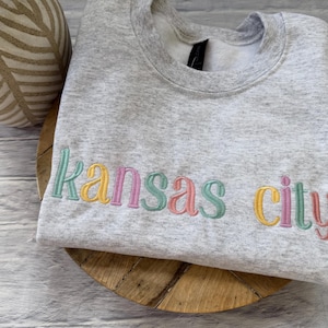 May include: A light gray sweatshirt with the embroidered text "Kansas City" in pastel colors. The sweatshirt is folded and rests on a wooden surface, with a decorative vase in the background.
