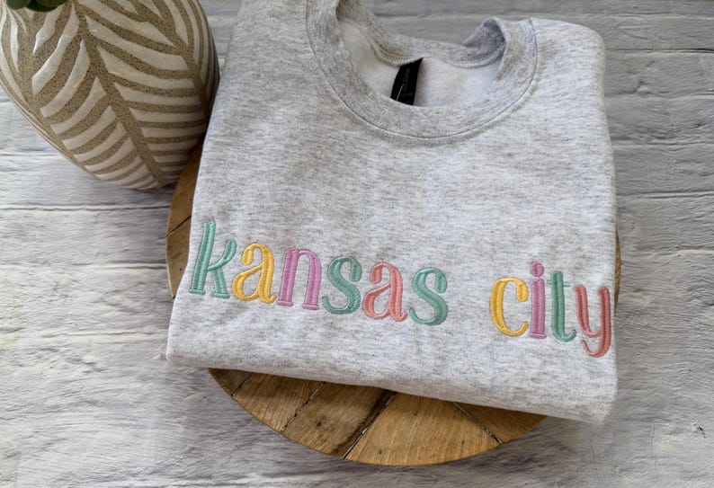 May include: A light gray sweatshirt with the embroidered text "Kansas City" in pastel colors. The sweatshirt is folded and rests on a wooden surface, with a decorative vase in the background.