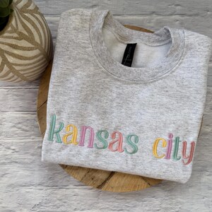 May include: A light gray sweatshirt with the embroidered text "Kansas City" in pastel colors. The sweatshirt is folded on a wooden surface, with a decorative vase in the background.