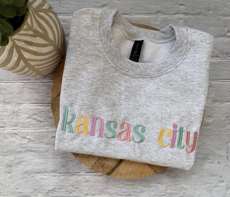 May include: A light gray sweatshirt with the embroidered text "Kansas City" in pastel colors. The sweatshirt is folded on a wooden surface, with a decorative vase in the background.