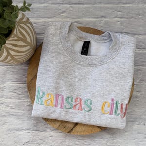 May include: A light gray sweatshirt with the embroidered text "Kansas City" in pastel colors. The sweatshirt is folded on a wooden surface, with a decorative plant in the background.