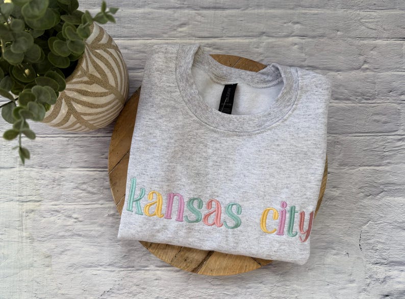 May include: A light gray sweatshirt with the embroidered text "Kansas City" in pastel colors. The sweatshirt is folded on a wooden surface, with a decorative plant in the background.
