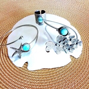 May include: A silver-toned jewelry set consisting of two open-ended bracelets and one wide band ring. Each piece features a round, light blue stone. One bracelet has a starfish motif, the other has a floral design, and the ring has a textured surface.