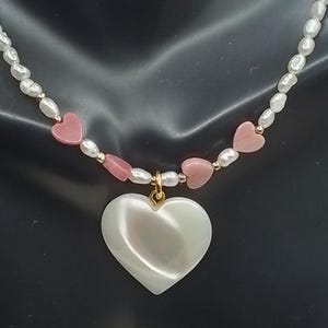 May include: A necklace featuring a large, pearlescent heart pendant suspended from a strand of white pearls and pink heart-shaped beads. The necklace is displayed on a black mannequin.