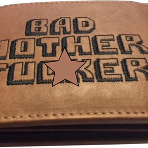 May include: A brown leather wallet featuring the text "BAD MOTHER FUCKER" in black, block lettering. A brown star is positioned between the words. The wallet has a stitched edge.