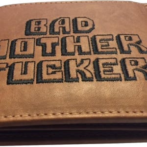 May include: A tan bi-fold wallet featuring the text BAD MOTHER FUCKER embroidered in black thread on the front exterior. The wallet has visible stitching along the edges and a smooth, matte finish across the surface of the brown material.