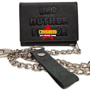 Pode incluir: Carteira de couro preta com texto em relevo "BAD MOTHER F CENSORED ER" e uma estrela vermelha. Inclui uma corrente prateada e uma tira de couro preta. O texto "SAYS THE REAL THANG" também é visível.