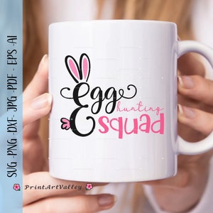 Könnte beinhalten: Eine weiße Keramiktasse mit einem schwarzen und rosafarbenen Design. Das Design zeigt eine Silhouette von Hasenohren und den Text "Egg Hunting Squad".