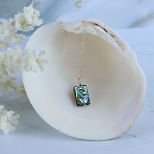 May include: A gold-toned chain necklace featuring a rectangular pendant made of iridescent abalone shell. The pendant displays swirling patterns of green, blue, and purple hues, suspended from a delicate metal link chain with a simple jump ring.