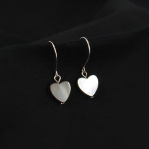 May include: A pair of heart-shaped earrings with a pearlescent finish. The earrings are suspended from silver-colored hooks, and the hearts have a subtle, iridescent sheen. The background is a solid black fabric.