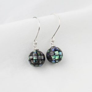 May include: A pair of silver dangle earrings with black and iridescent round beads. The beads are made of abalone shell and have a mosaic pattern.