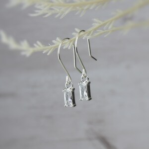 May include: A pair of silver-toned drop earrings. Each earring features a rectangular, clear gemstone set in a silver frame. The earrings are suspended from curved ear wires, and are displayed against a soft, blurred background.