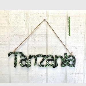 May include: Wooden sign spelling "Tanzania" in a bold, stylized font. The letters are covered in a black and green patterned fabric. The sign hangs from a natural twine string against a white, textured background.
