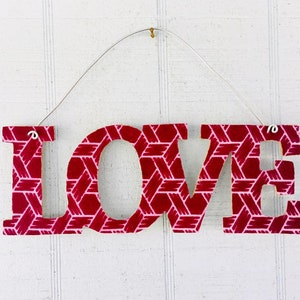 Könnte beinhalten: Ein hängendes Wandschild mit dem Wort LOVE in Blockbuchstaben. Die Buchstaben haben ein gewebtes Korbgeflechtmuster in Dunkelrot und Hellrosa. Ein dünner silberner Draht ist oben am L und E befestigt, um das Schild an die Wand zu hängen.