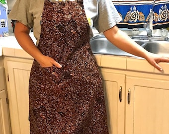 Könnte beinhalten: Eine braune Schürze mit einem komplizierten, sich wiederholenden Paisley- und Blumenmuster in Dunkelbraun, Kupfer und Schwarz. Die Schürze verfügt über eine einzelne Vordertasche auf der rechten Seite, ein Nackenband und Taillenbänder.