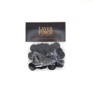 May include: A clear plastic bag filled with black, circular game tokens. The bag has a black label with gold text that reads "LAYER FORGE STUDIO" and the website address. The tokens appear to be made of plastic and have a detailed design.
