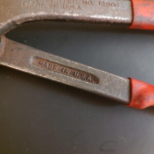 May include: A pair of metal bolt cutters with red rubberized handle grips. The tool features embossed text on the metal arms that reads H.K. PORTER NO. 12906 MADE IN U.S.A. and a secondary stamp on the lower arm that reads MADE IN U.S.A.