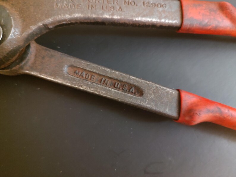 May include: A pair of metal bolt cutters with red rubberized handle grips. The tool features embossed text on the metal arms that reads H.K. PORTER NO. 12906 MADE IN U.S.A. and a secondary stamp on the lower arm that reads MADE IN U.S.A.
