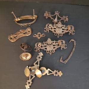 May include: A collection of assorted antique-style metal furniture hardware including several ornate drawer pulls, a keyhole escutcheon plate, a ring pull, and a decorative handle, all featuring a weathered brass or bronze finish.