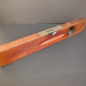 May include: A vintage wooden spirit level featuring a rectangular body with a warm brown finish. The tool includes a central metal plate housing a horizontal glass vial and a circular glass vial set into the wood for measuring level surfaces.