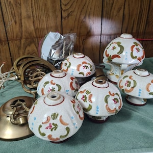 May include: A collection of five off-white ceramic light fixture shades featuring a hand-painted floral pattern in green, orange, and blue. The set includes brass-colored metal hardware components, including a circular ceiling canopy and curved arms.