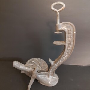 May include: A vintage cast iron Enterprise Philadelphia food press featuring a vertical clamp mechanism with a threaded screw handle and a side-mounted hand crank attached to a circular wheel. The metal surface shows a weathered grey patina.