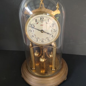 May include: A gold-toned anniversary clock with a glass dome cover, featuring a cream-colored face with black Arabic numerals, floral accents, and the brand name Kundo. The clock has a brass base, three support pillars, and a rotating pendulum.