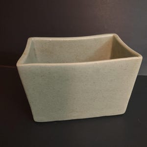 May include: A rectangular ceramic planter with a light beige, speckled finish. The vessel features slightly flared sides and a smooth, matte texture, with a wide open top and a flat base, presenting a clean and simple design for holding items.