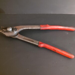May include: A pair of metal fencing pliers featuring a silver-toned steel construction with a pivot point and two long handles covered in worn red rubber grips. The tool has a pointed jaw design with a small cutting edge and a central hinge bolt.