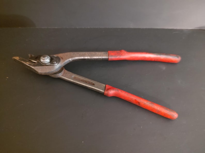 May include: A pair of metal fencing pliers featuring a silver-toned steel construction with a pivot point and two long handles covered in worn red rubber grips. The tool has a pointed jaw design with a small cutting edge and a central hinge bolt.