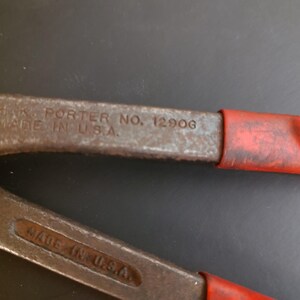 May include: A pair of metal bolt cutters with red rubberized handles. The tool features embossed text on the metal arms that reads H.K. PORTER NO. 1290G and MADE IN U.S.A. The metal surface shows signs of wear and oxidation, with a dark grey finish.