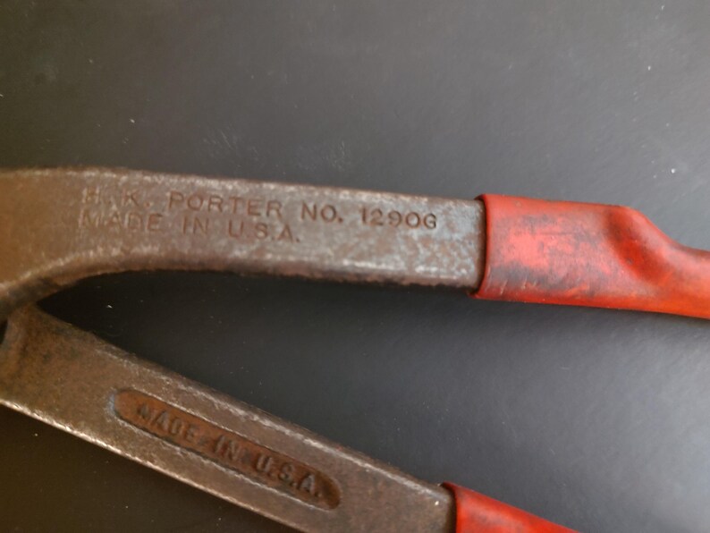 May include: A pair of metal bolt cutters with red rubberized handles. The tool features embossed text on the metal arms that reads H.K. PORTER NO. 1290G and MADE IN U.S.A. The metal surface shows signs of wear and oxidation, with a dark grey finish.