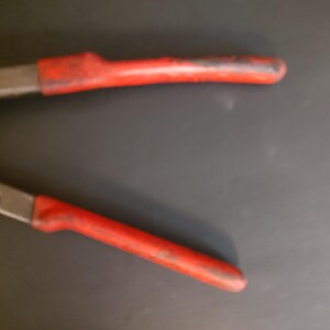 May include: A pair of metal pliers featuring long, slender jaws and handles coated in a worn, textured red rubber grip. The metal components show a dull, weathered finish, while the red handles exhibit signs of use with visible scuffs and dark marks along the surface, indicating a tool designed for gripping or bending tasks.
