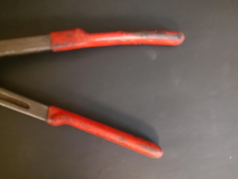 May include: A pair of metal pliers featuring long, slender jaws and handles coated in a worn, textured red rubber grip. The metal components show a dull, weathered finish, while the red handles exhibit signs of use with visible scuffs and dark marks along the surface, indicating a tool designed for gripping or bending tasks.