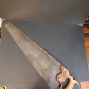 May include: A vintage hand saw featuring a long, tapered steel blade with a serrated edge and a contoured wooden handle. The handle is secured to the blade with three metal rivets and features a circular metal medallion embedded in the wood.