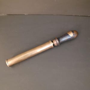 May include: A vintage brass syringe featuring a long, cylindrical barrel and a dark, weathered wooden handle. The metal surface shows signs of age and patina, with a distinct transition point between the brass body and the handle attachment.