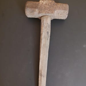 May include: A heavy-duty sledgehammer featuring a large, rectangular, weathered metal head with a textured, rusted surface and a long, straight, light-colored wooden handle that shows visible grain and signs of wear along its length.