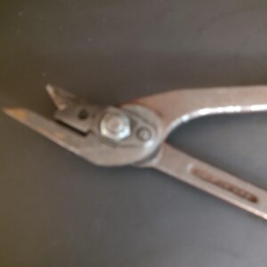 May include: A pair of metal aviation snips featuring sharp, angled cutting blades, a central pivot bolt, and two long handles with red rubberized grips. The tool is constructed from durable steel with a matte finish, designed for cutting sheet metal.