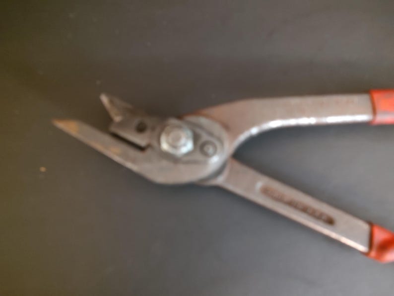 May include: A pair of metal aviation snips featuring sharp, angled cutting blades, a central pivot bolt, and two long handles with red rubberized grips. The tool is constructed from durable steel with a matte finish, designed for cutting sheet metal.