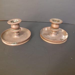 May include: A pair of matching brass candle holders, each featuring a wide, circular drip tray base and a central stem with a rounded, tiered design. The metal surface displays a warm, metallic tone with subtle signs of age and patina.