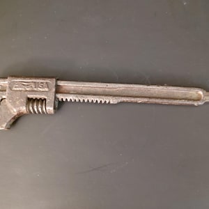May include: A vintage metal pipe wrench with a textured, dark grey finish. The tool features an adjustable jaw mechanism with a threaded nut and a long, straight handle. The metal surface is embossed with the text USA near the head.