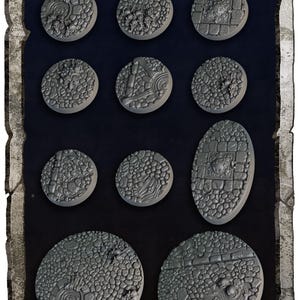 May include: A collection of gray, textured miniature bases in various shapes, including round and oval. The bases feature detailed, sculpted designs resembling stone, terrain, and architectural elements, suitable for tabletop gaming.