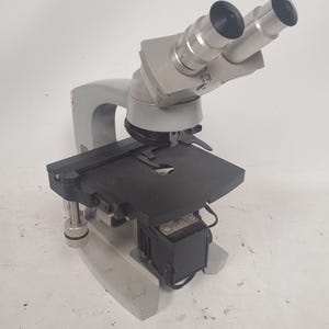 May include: A light gray microscope with two eyepieces. The microscope has a black stage and a light gray base. The eyepieces are silver and black. The microscope is designed for scientific or educational use.