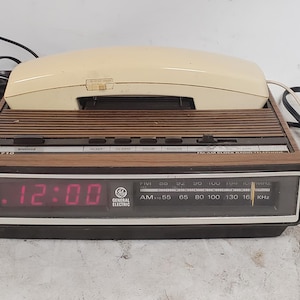 Puede incluir: Radio reloj digital General Electric vintage con un auricular telefónico color crema. El reloj muestra las 12:00 en dígitos rojos. La radio tiene un acabado de veta de madera y un dial de radio AM.
